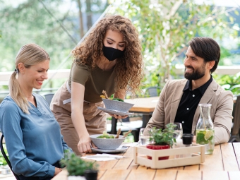 Waitress with face mask serving happy couple outdoors on terrace restaurant