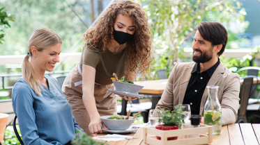 Waitress with face mask serving happy couple outdoors on terrace restaurant