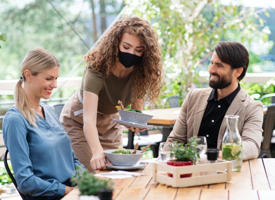 Waitress with face mask serving happy couple outdoors on terrace restaurant