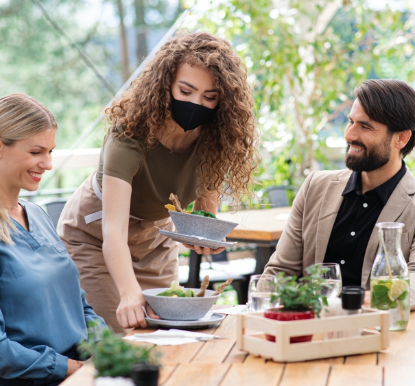 Waitress with face mask serving happy couple outdoors on terrace restaurant